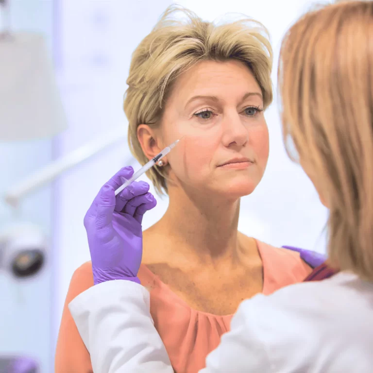 Žena s krátkými blond vlasy podstupující injekční aplikaci botulotoxinu do oblasti spánku od lékaře v rukavicích.