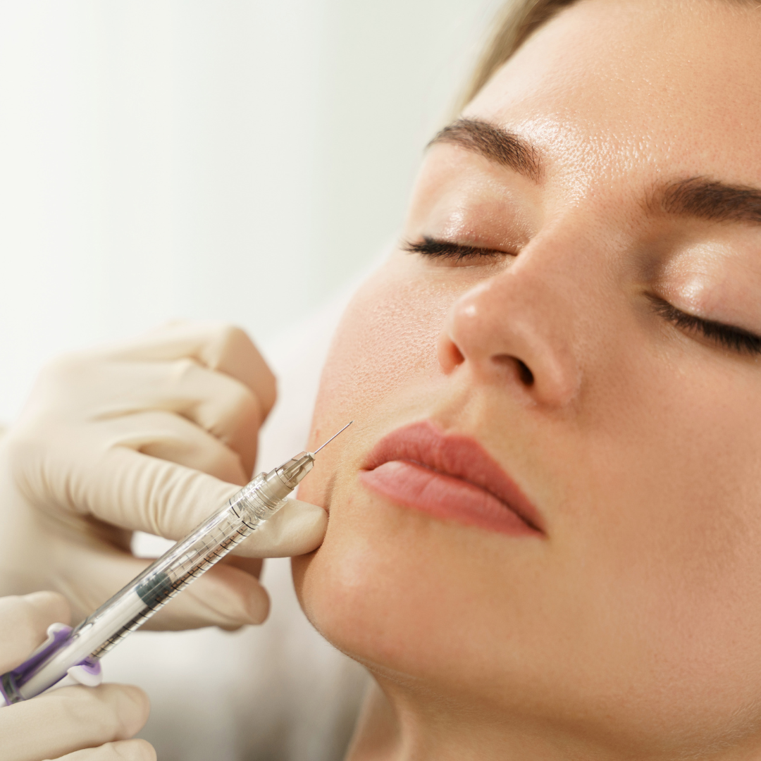 Žena během kosmetické procedury, kdy ruka v rukavici aplikuje injekci s kyselinou polymléčnou do oblasti tváře.